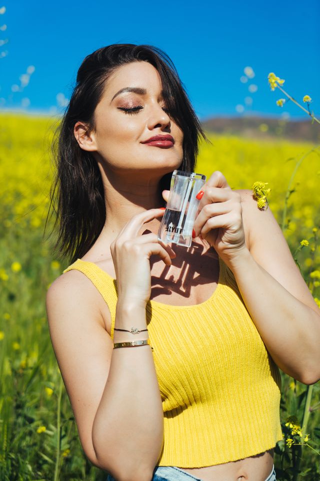 Woman in a yellow top using a RYOT dugout in a flower fieldWoman in a yellow top holding a vintage camera to her face while standing in a yellow flower field under a blue sky